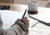 person writing on brown wooden table near white ceramic mug